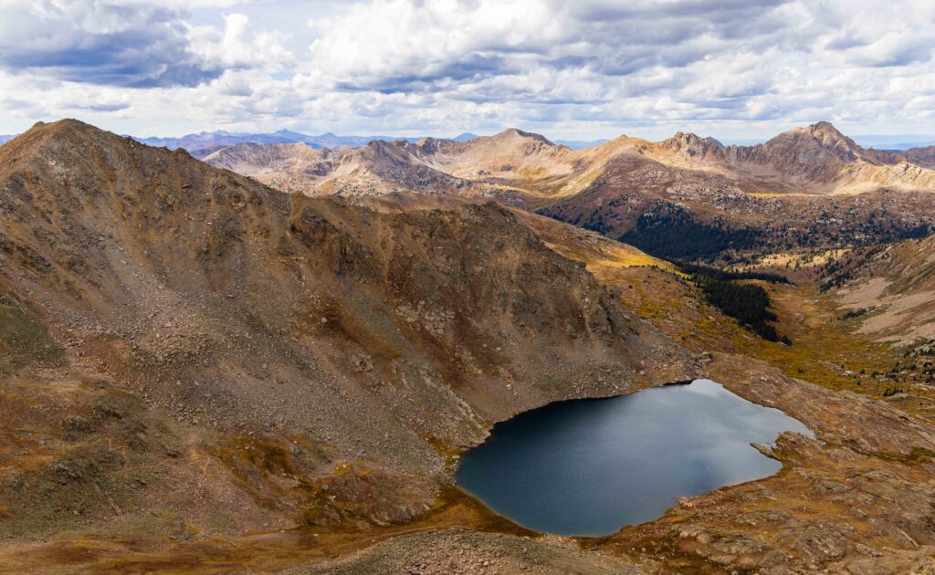 Aerial view of a dark alpine lake below a rocky ridge with a mountain range beyond