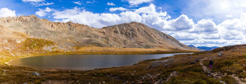 Panoramic view of an alpine lake with a hiker on the shoreline trail below rocky peaks