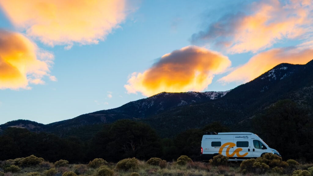 a campervan sits on the side of the road with colorful clouds and mountains above