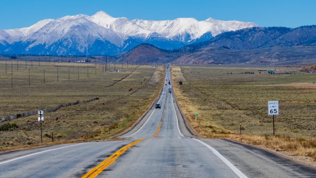 a straight road with cars on it and snowy mountains in the distance