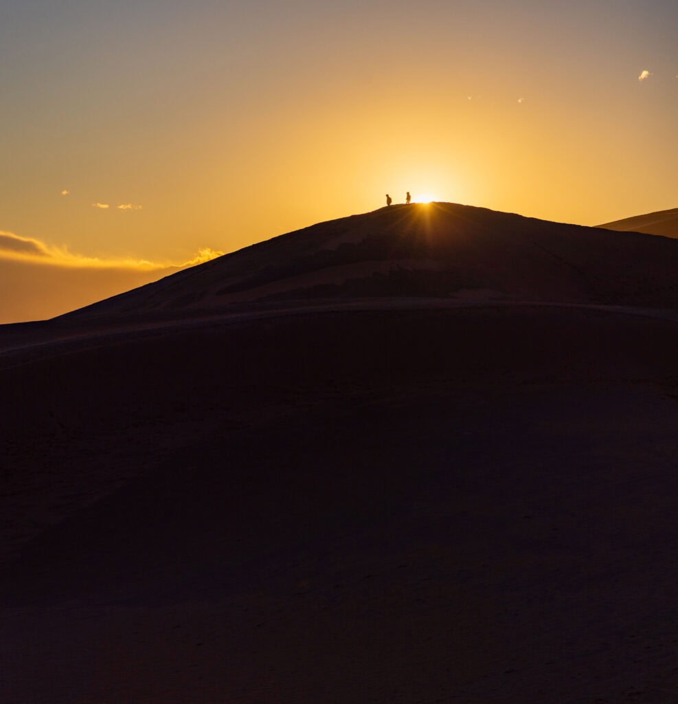 two hikers on top of a dune with the sun behind them