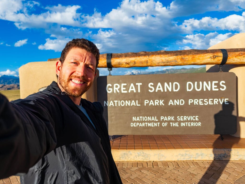 a visitor smiles in front of the great sand dunes national park entrance sign